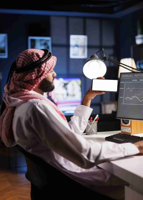 Side-view shot Middle Eastern man in traditional attire sitting at an office desk, using a desktop computer. They hold a mobile phone with a blank chromakey mockup template, offering space for advertising.