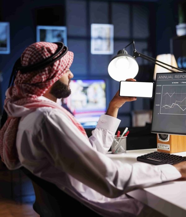 Side-view shot Middle Eastern man in traditional attire sitting at an office desk, using a desktop computer. They hold a mobile phone with a blank chromakey mockup template, offering space for advertising.